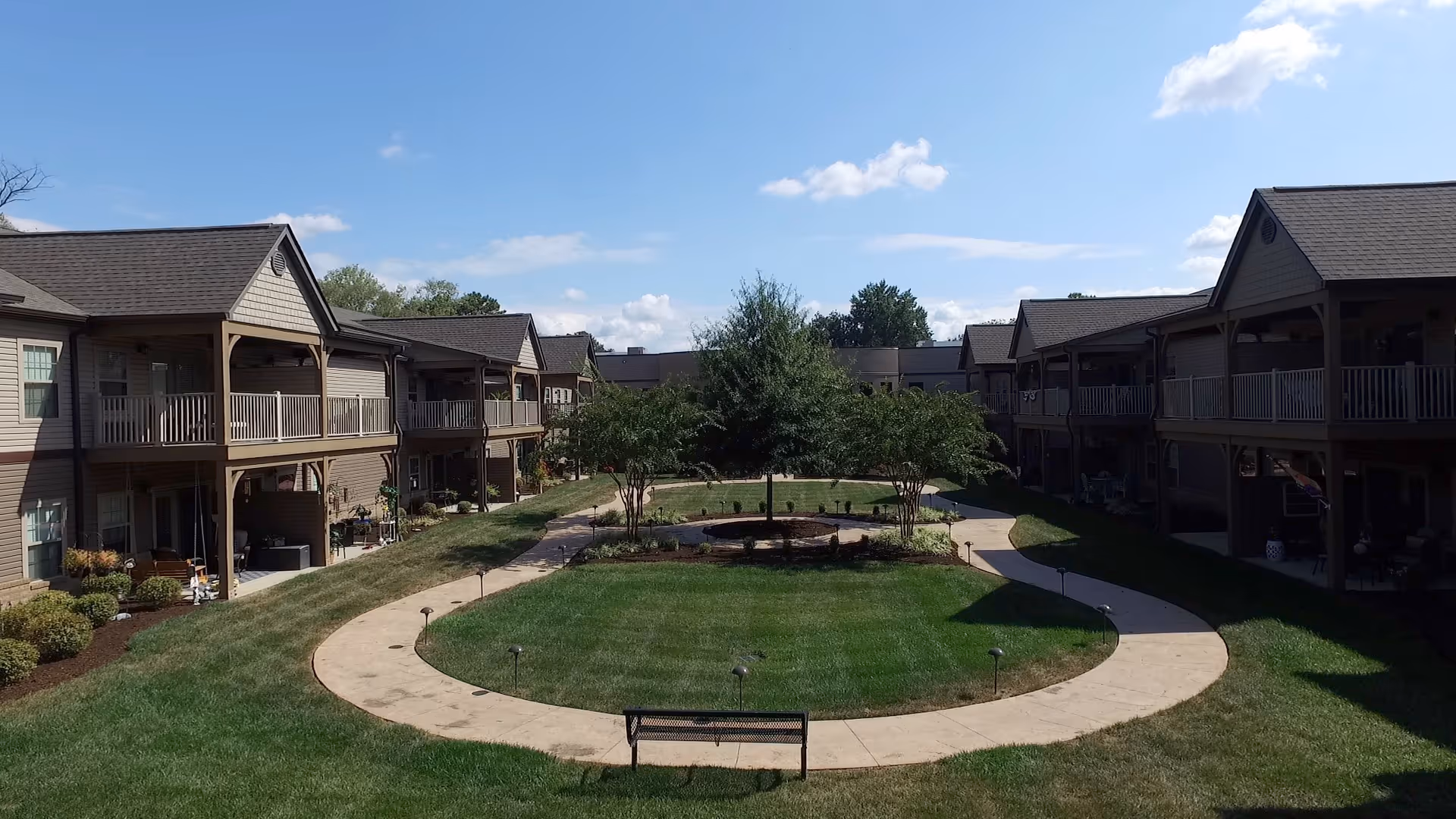 Outdoor courtyard area at Parkview Senior Living - West Knoxville featuring a circular walking path surrounding a grassy area with trees and a bench, flanked by two-story residential buildings with balconies under a clear blue sky.