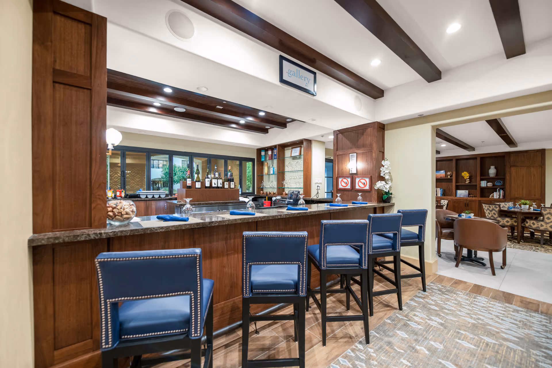 Interior view of a bar area with a marble countertop and five blue cushioned bar stools. Behind the bar, there are bottles of wine and glasses on shelves. The ceiling has wooden beams and recessed lighting. Adjacent to the bar is a seating area with tables and chairs, and wooden bookshelves with decorative items.
