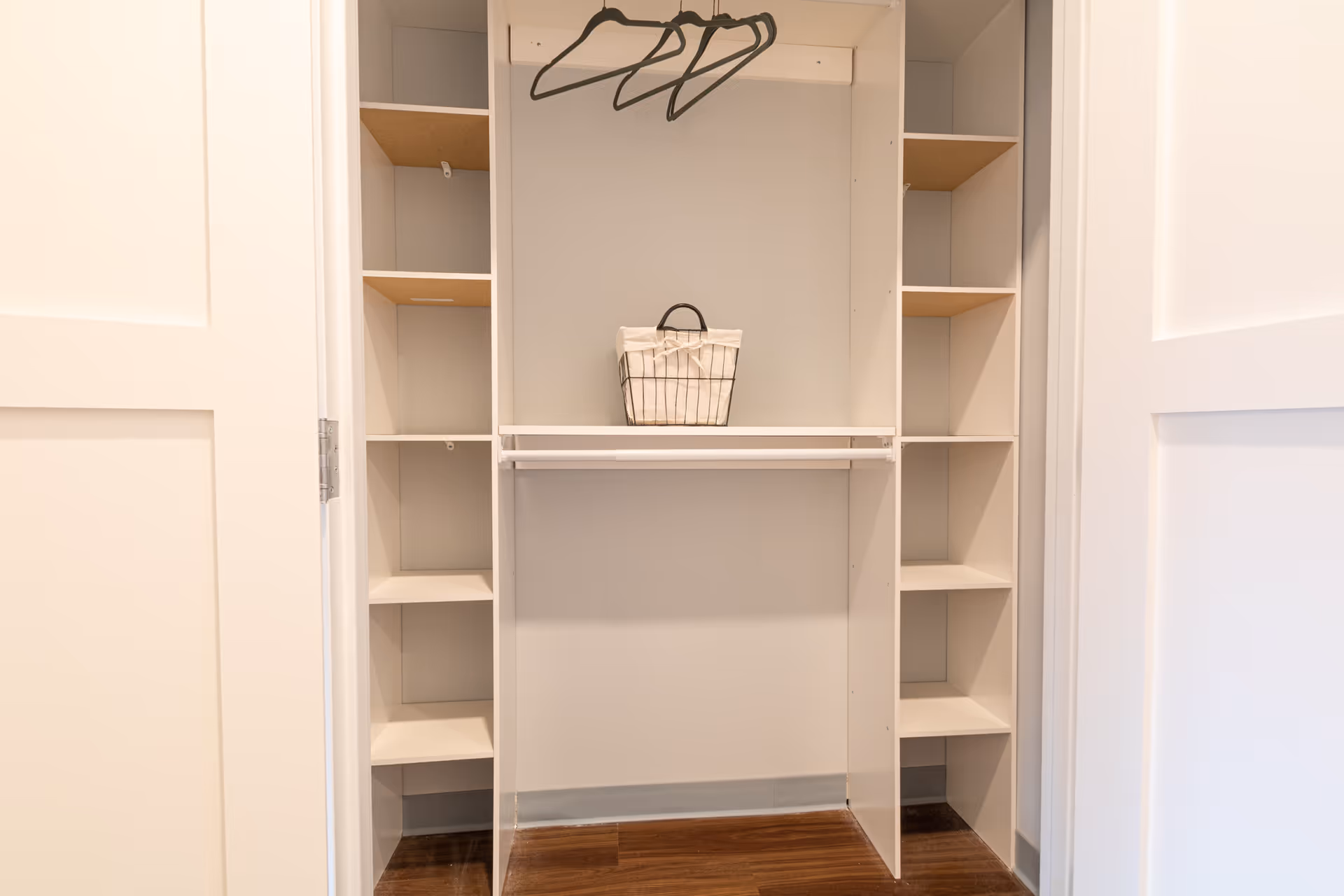 An empty white closet with multiple shelves on both sides, a hanging rod with three black hangers, and a small basket with a fabric liner placed on the middle shelf. The closet doors are open, revealing a wooden floor beneath.