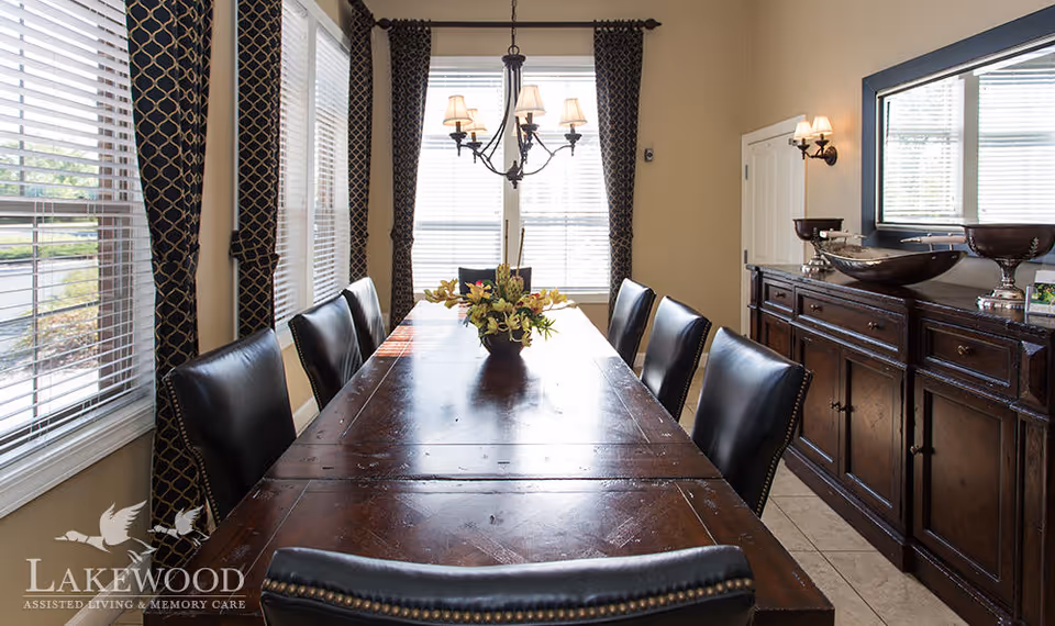 Long wooden dining table with leather chairs, a floral centerpiece, chandelier, sideboard, and windowed walls with curtains.