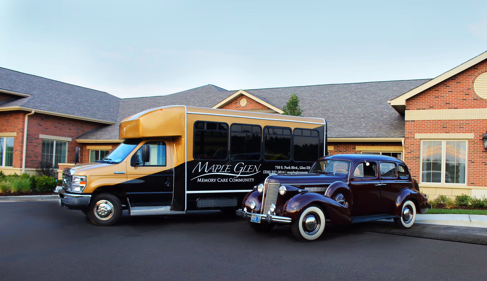 A shuttle bus and a vintage car parked in front of a brick building with multiple windows and a gray roof. The shuttle bus has signage for Maple Glen Memory Care Community, including an address and phone number.