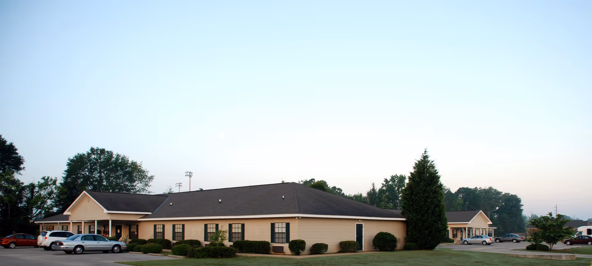 Exterior view of a single-story senior living facility building with beige siding and a dark roof, surrounded by a well-maintained lawn and several parked cars in the parking lot. Trees and a clear sky are visible in the background.