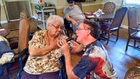 An elderly woman in a wheelchair is holding a microphone and appears to be singing or speaking with a man kneeling in front of her who is also holding the microphone. Another elderly man in a wheelchair is seated at a table in the background in a room with wooden floors and chairs around tables.
