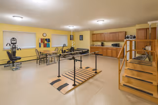 A rehabilitation room with exercise equipment including a stationary bike, parallel bars for walking practice, a rowing machine, and a set of stairs for physical therapy. The room has yellow walls, beige flooring, wooden cabinets along one wall, and windows with blinds.