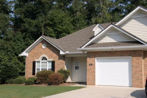 Exterior view of a single-story brick and siding residential building with a white garage door, a window with black shutters, and a small front yard with bushes and grass. Trees are visible in the background.