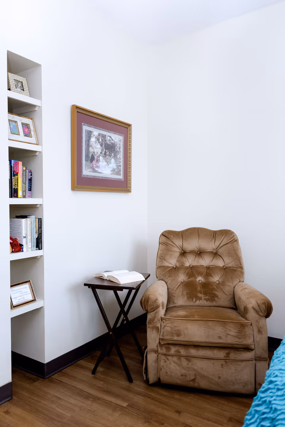 A brown upholstered recliner beside a small folding side table with an open book, built-in bookshelf and framed artwork on a white wall.