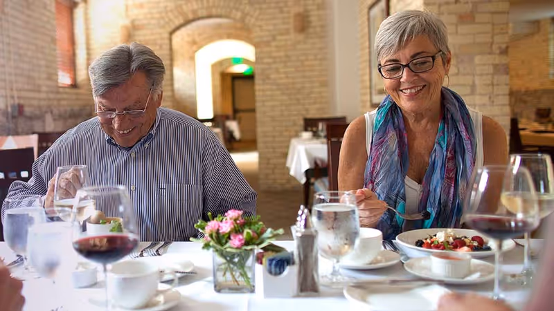 An elderly man and woman sitting at a dining table in a restaurant, enjoying their meal. The table is set with glasses of water, wine, cups, and a small flower arrangement. The background shows a warm, inviting interior with brick walls and arched doorways.