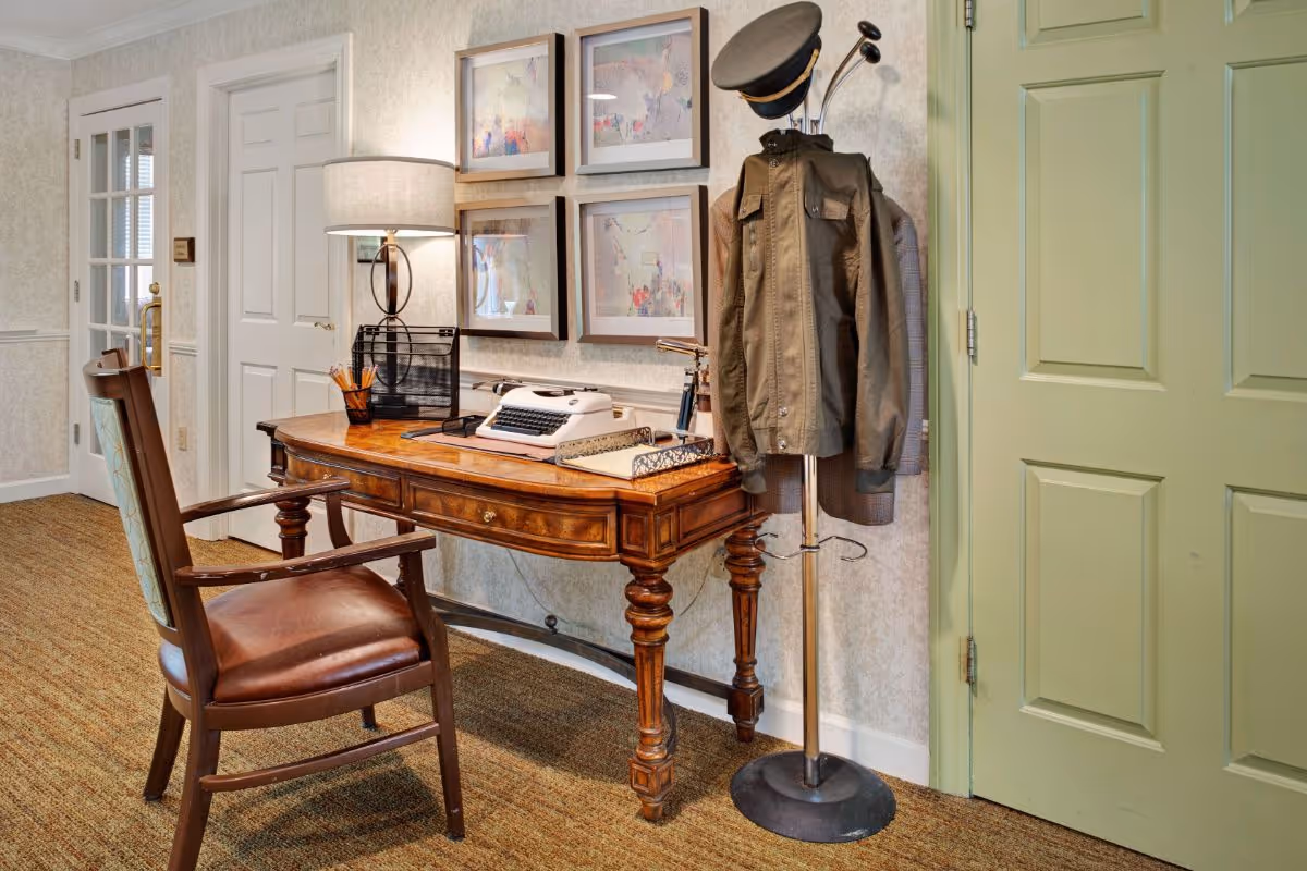 A cozy interior corner featuring a wooden desk with a vintage typewriter, a lamp, and a pencil holder. A wooden chair with a cushioned seat is placed in front of the desk. On the right side, there is a coat rack holding a jacket and a hat. The walls are decorated with four framed abstract paintings, and there are two doors, one white and one light green.
