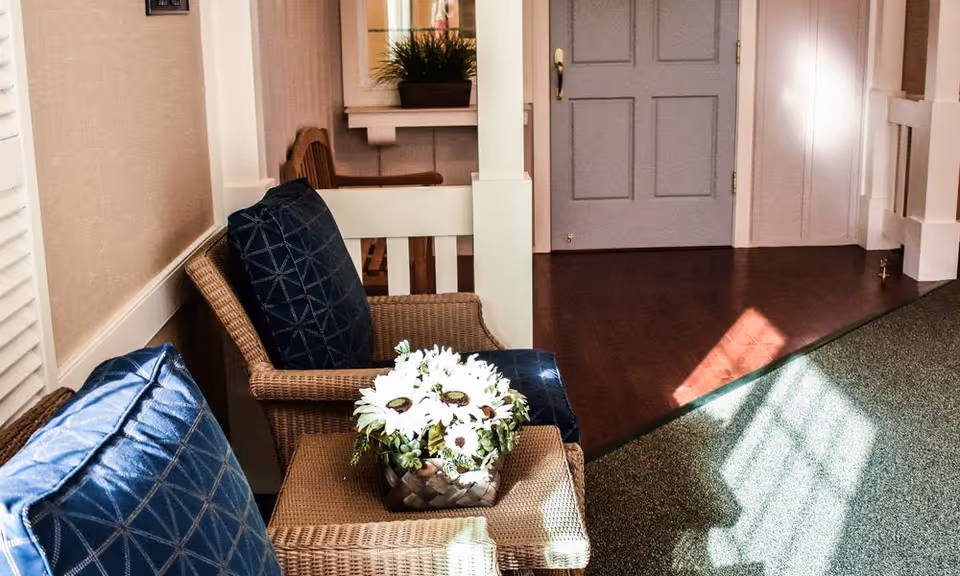 A cozy seating area with two wicker chairs featuring blue cushions and a small wicker table between them holding a flower arrangement with white daisies. The area is well-lit with sunlight casting shadows on the carpeted floor. In the background, there is a gray door and a small window with a plant on the sill.