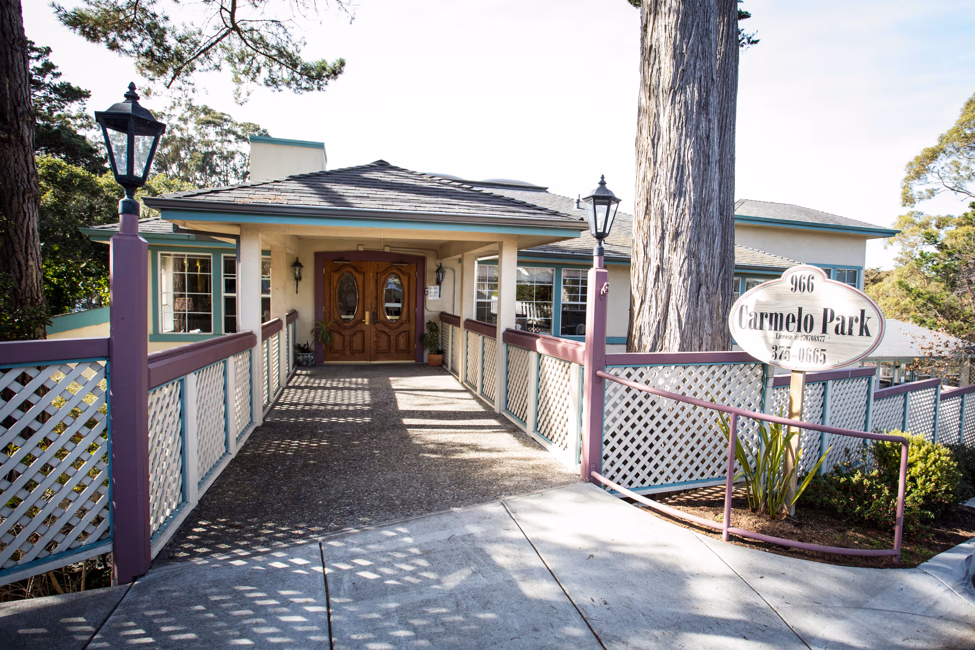 Entrance to Carmelo Park facility with a covered walkway leading to double wooden doors. The walkway has white lattice fencing with purple railings and two black lamp posts on either side. A sign near the entrance displays the name Carmelo Park and a phone number. Trees and greenery surround the building.