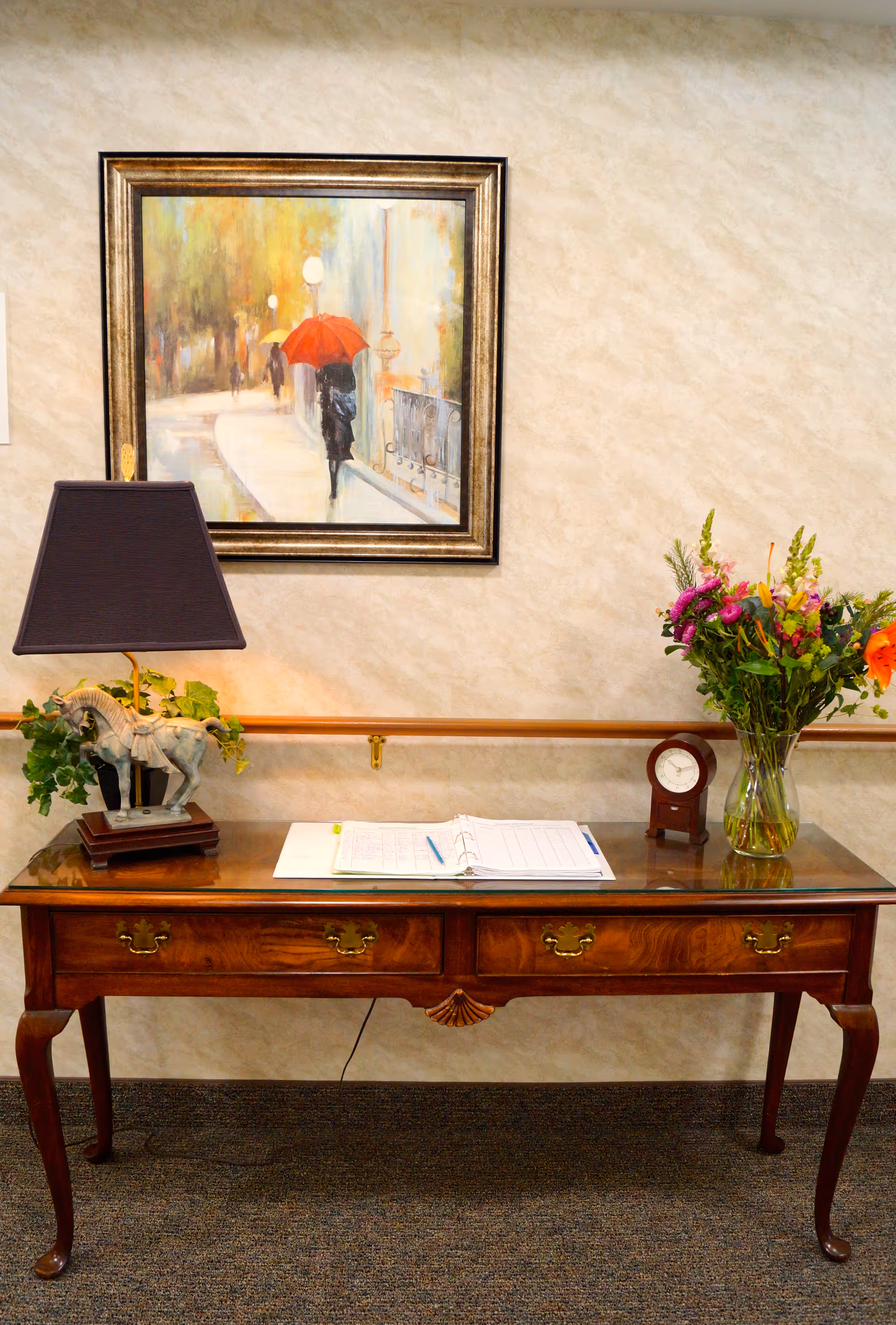 A wooden table with a glass top against a beige textured wall. On the table, there is a decorative lamp with a horse figurine base, an open binder with papers and a pen, a small round clock, and a vase with colorful flowers. Above the table hangs a framed painting of a person walking with a red umbrella on a rainy street.