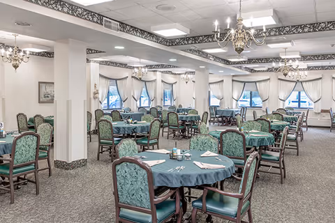 A spacious dining room with multiple round tables covered with teal tablecloths, each surrounded by green upholstered chairs. The room features large windows with white curtains, chandeliers hanging from the ceiling, and a patterned carpeted floor.