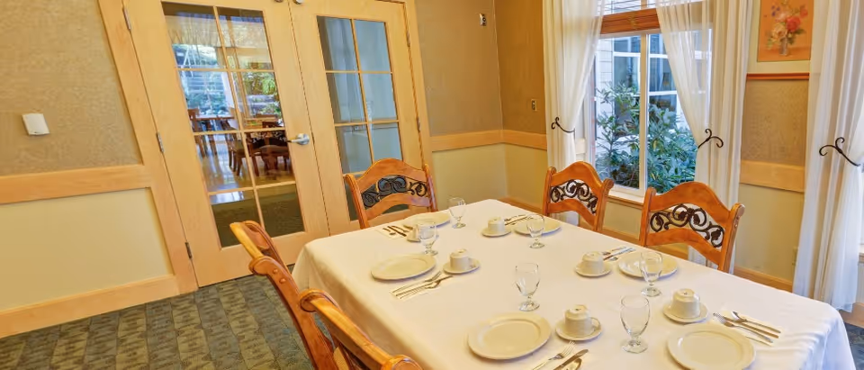A dining room table set for six with white tablecloth, plates, cups, glasses, and silverware. The room has wooden chairs with decorative backs, beige walls with wooden trim, a window with white curtains, and double glass doors leading to another room.