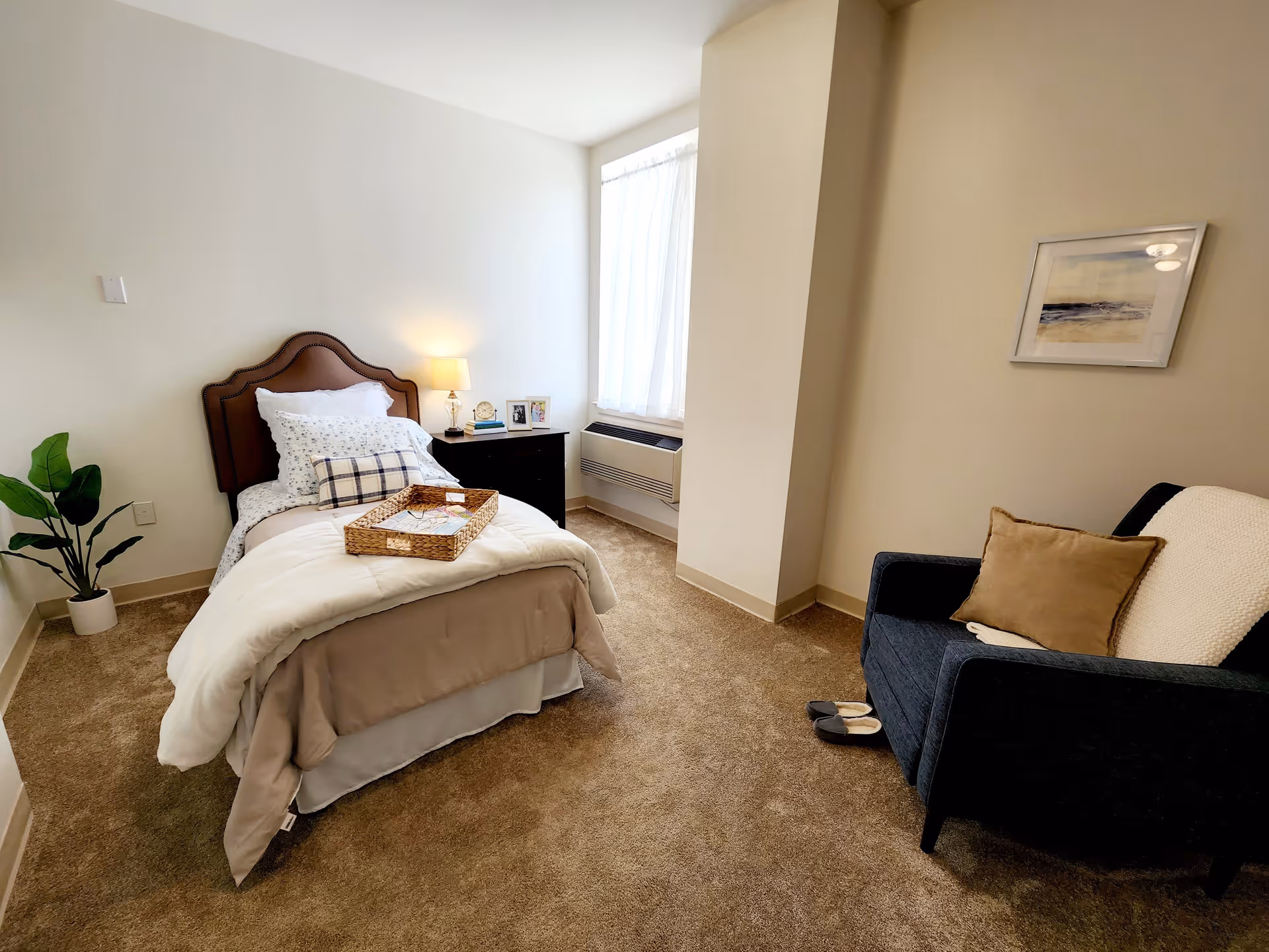 A cozy bedroom with a single bed featuring a brown upholstered headboard, white and beige bedding, and a wicker tray on top. Next to the bed is a dark wooden nightstand with a lamp, clock, books, and framed photos. A large window with sheer white curtains lets in natural light. Across from the bed is a dark blue armchair with a beige pillow and a white throw blanket, with a pair of slippers on the carpeted floor. A framed picture hangs on the wall above the chair.