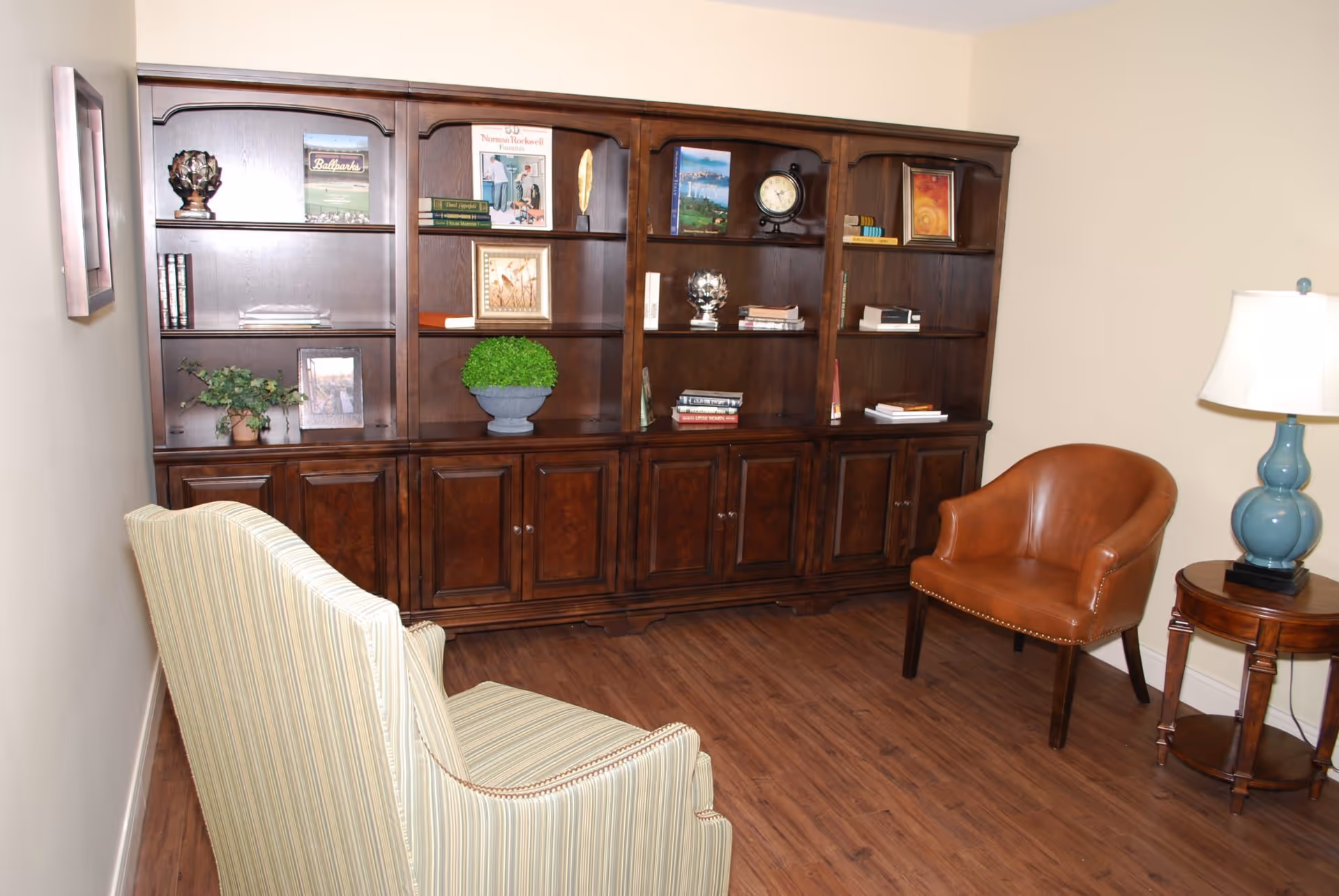 A cozy interior room featuring a large wooden bookshelf filled with books and decorative items, a striped upholstered armchair, a brown leather chair, a small wooden side table with a blue lamp, and wood flooring.