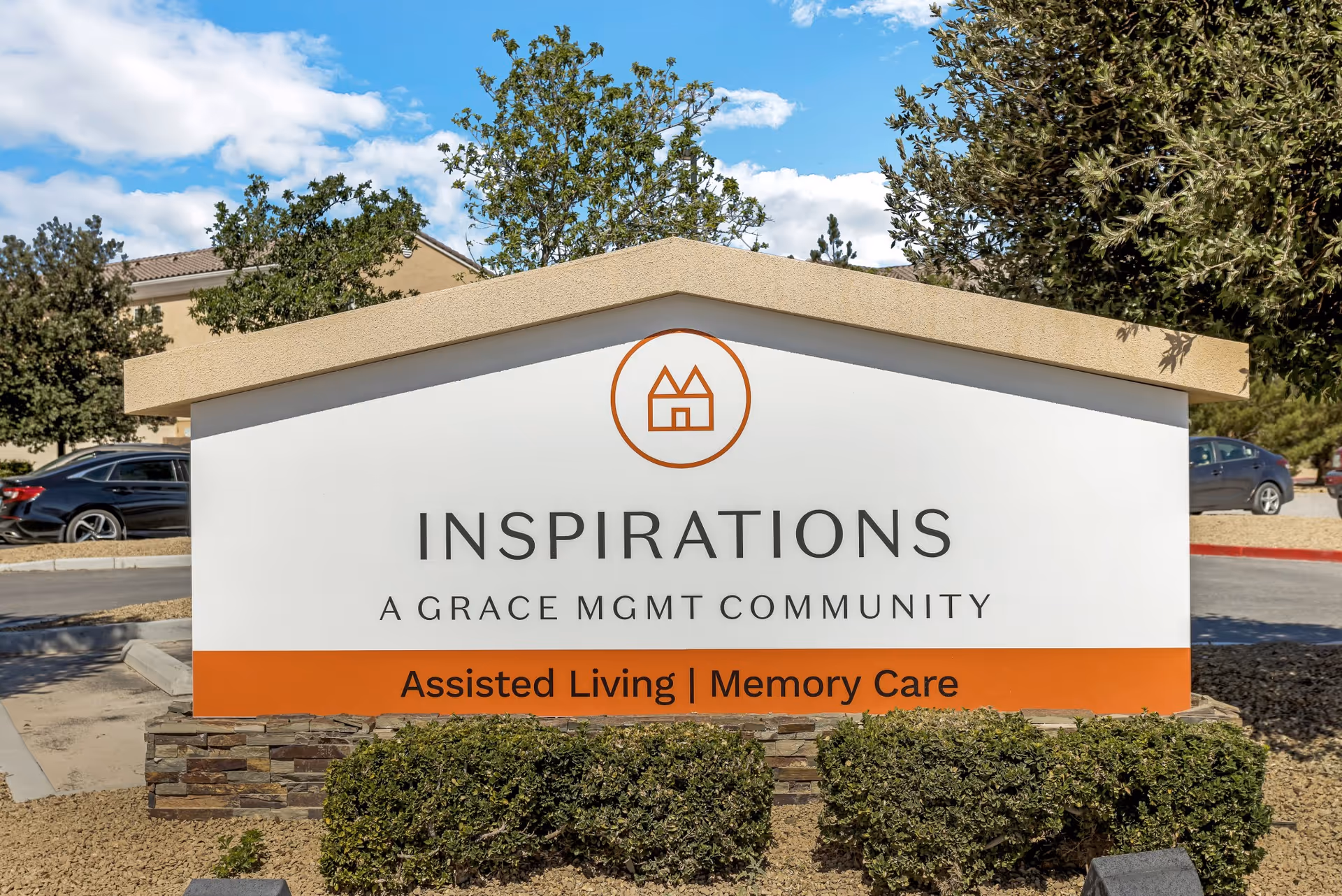 Outdoor sign for Inspirations, a Grace Management community offering Assisted Living and Memory Care, with cars and trees in the background under a blue sky with clouds.