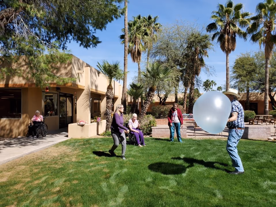 A group of elderly people and a caregiver are enjoying an outdoor activity on a grassy lawn at Woodland Palms Memory Care. One man is holding a large white balloon, while others watch and participate. The setting includes palm trees, a beige building, and clear blue sky.