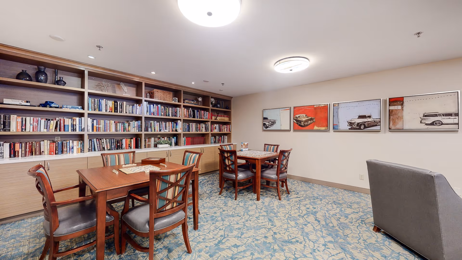 A cozy room with wooden bookshelves filled with books and decorative items along one wall. There are two wooden tables with four chairs each, arranged for reading or playing games. The carpet has a blue and beige pattern, and the walls are decorated with framed artwork featuring vintage cars. A gray armchair is positioned near the right side of the room, and the ceiling has modern light fixtures.
