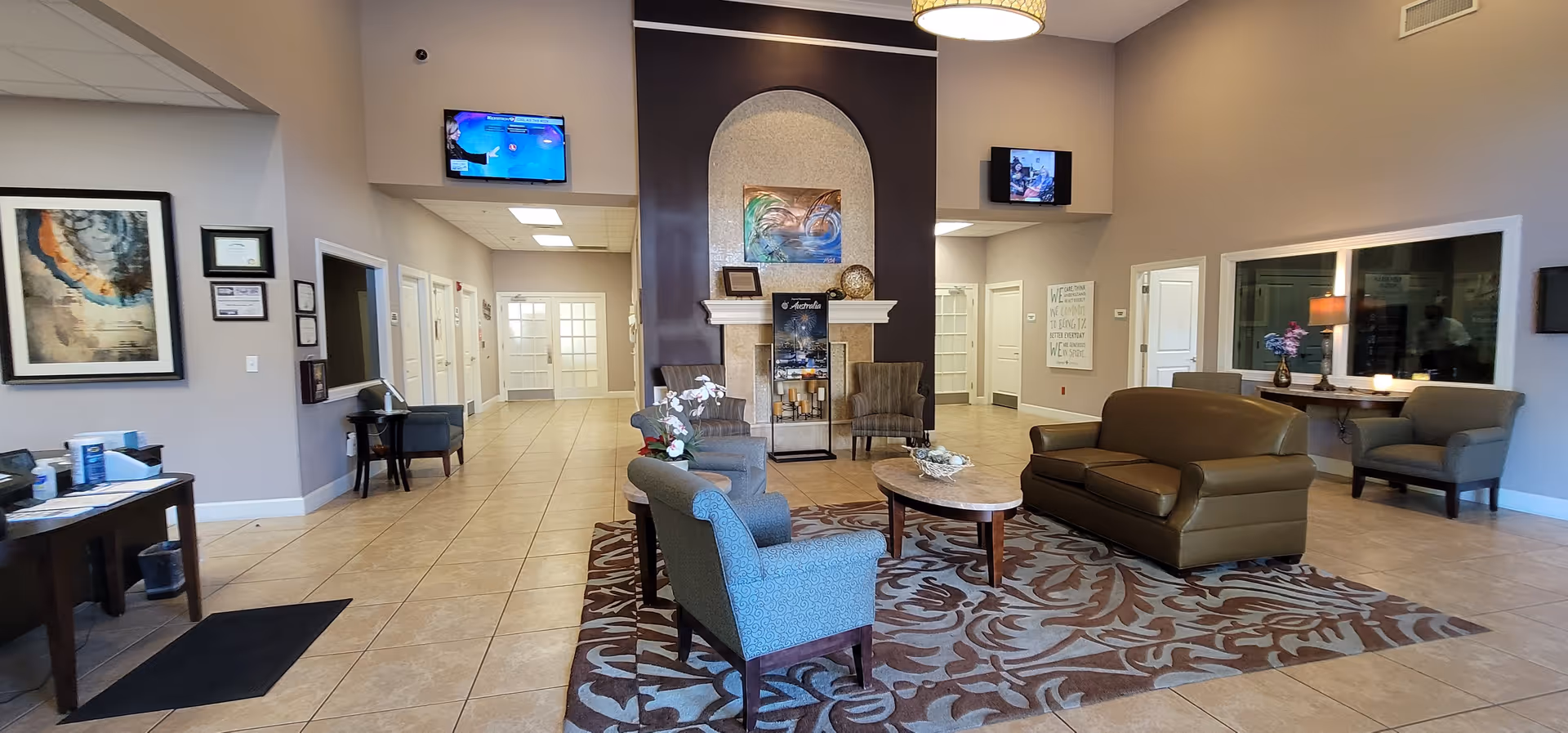 A spacious and well-lit common area in a senior living facility with a seating arrangement including armchairs and a sofa around a coffee table on a patterned rug. The room features a decorative fireplace with artwork above it, two wall-mounted televisions, and a hallway leading to other rooms. The walls are painted in neutral tones, and there are framed pictures and a console table with a lamp and flowers.