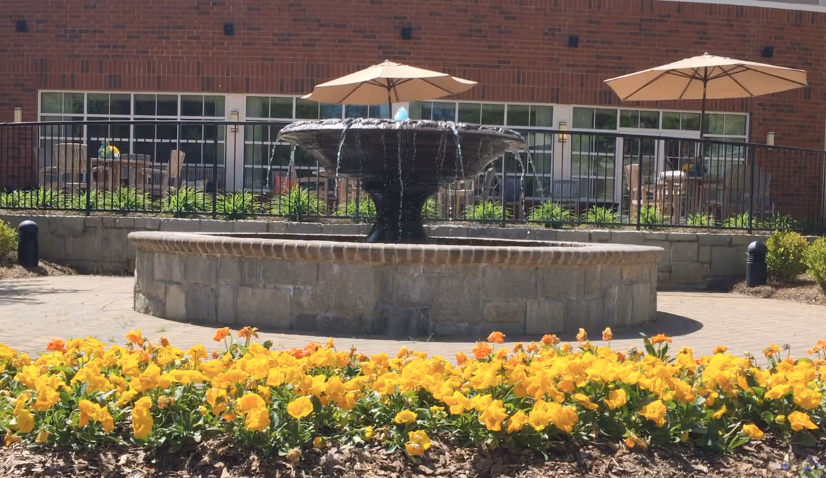 Outdoor courtyard area with a stone water fountain surrounded by a flower bed of yellow and orange flowers. Behind the fountain, there is a patio with tables, chairs, and umbrellas in front of a brick building with large windows.