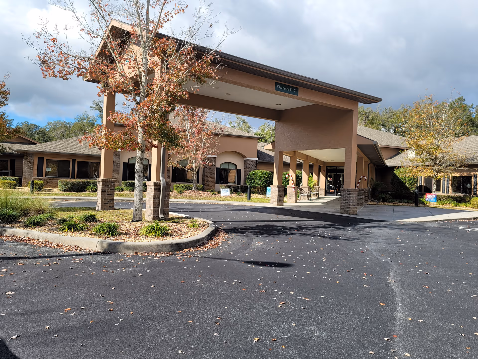 Exterior view of Best Care Senior Living at Lecanto facility showing the entrance with a covered drop-off area, surrounded by trees with autumn leaves and a paved driveway.