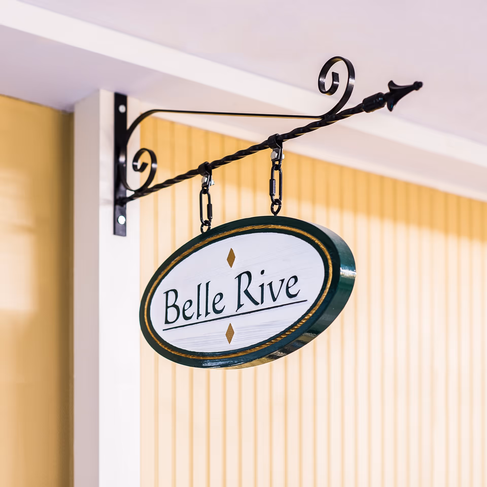 A decorative hanging sign with the text 'Belle Rive' mounted on a black wrought iron bracket against a yellow paneled wall.