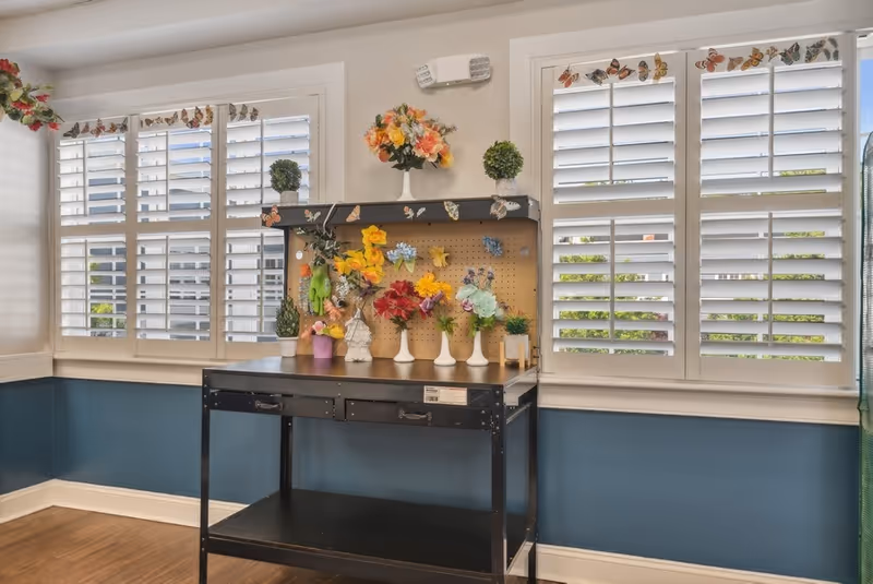 A bright room with two large windows featuring white plantation shutters decorated with butterfly stickers. Below the windows is a black metal table with a pegboard back, adorned with various colorful artificial flowers and small potted plants. The walls are painted white on top and blue on the bottom half, with a wooden floor.