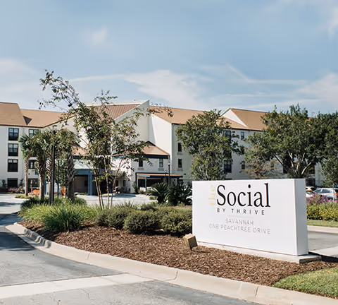 Exterior view of The Social at Savannah senior living facility with a large white sign in the foreground displaying the facility's name and address. The building is multi-story with beige walls and a brown roof, surrounded by trees and landscaping under a partly cloudy sky.