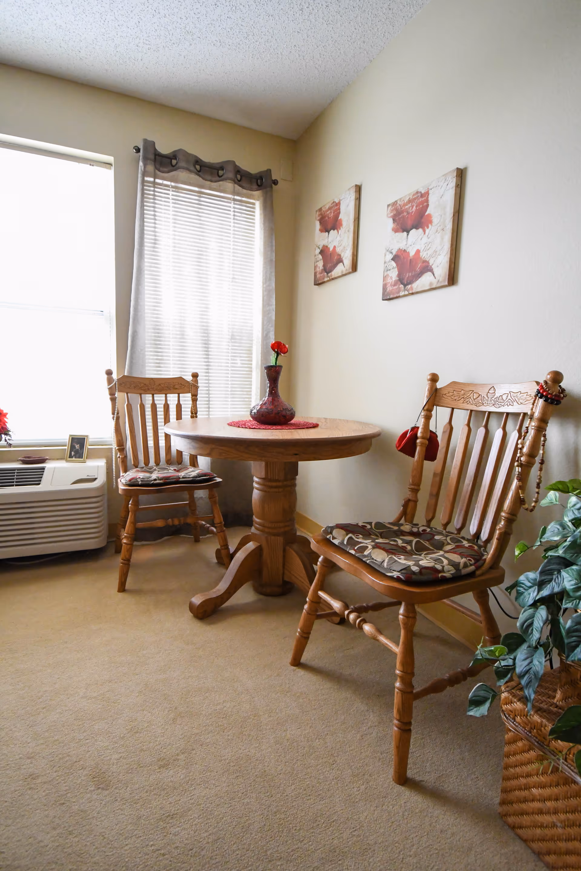 A small dining area with a round wooden table and two wooden chairs with patterned cushions. A red vase with a single red flower is on the table. The room has beige walls with two floral paintings, a window with sheer curtains, a wall-mounted air conditioning unit, and a green potted plant in a woven basket on the floor.