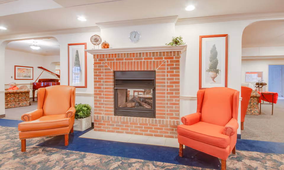 A cozy common area with a brick fireplace in the center, flanked by two orange upholstered armchairs. The fireplace mantel has decorative items including a clock and plants. The room has framed artwork on the walls and a patterned carpet. In the background, there is a grand piano and additional seating with floral upholstery.