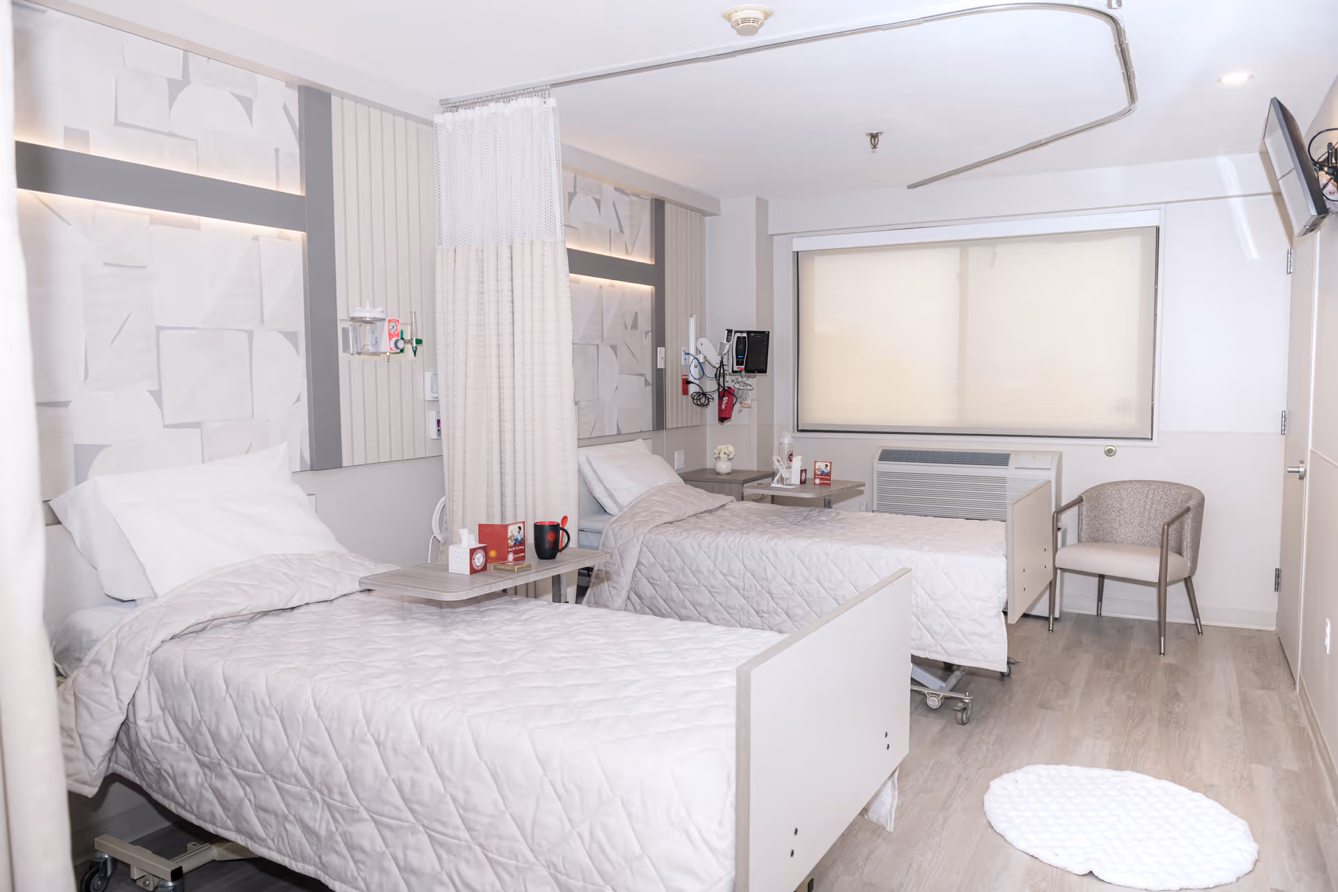 A clean and bright skilled nursing facility room with two hospital beds separated by a privacy curtain. Each bed has white bedding and pillows. There are bedside tables with personal items, medical equipment mounted on the wall, a chair near the window with closed blinds, and a wall-mounted television. The floor is light wood, and the walls are painted in neutral tones.