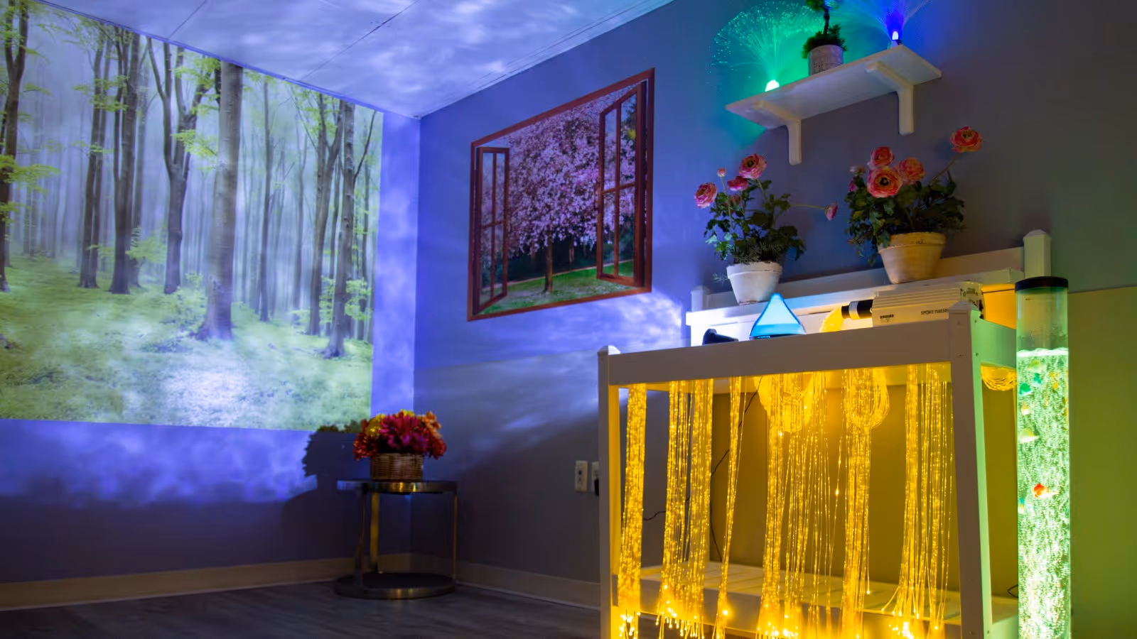 A sensory room with calming light effects including a forest scene projected on the wall, a faux window with a cherry blossom tree image, fiber optic lights hanging from a white frame, potted plants on shelves, and a cylindrical bubble tube light emitting green light.