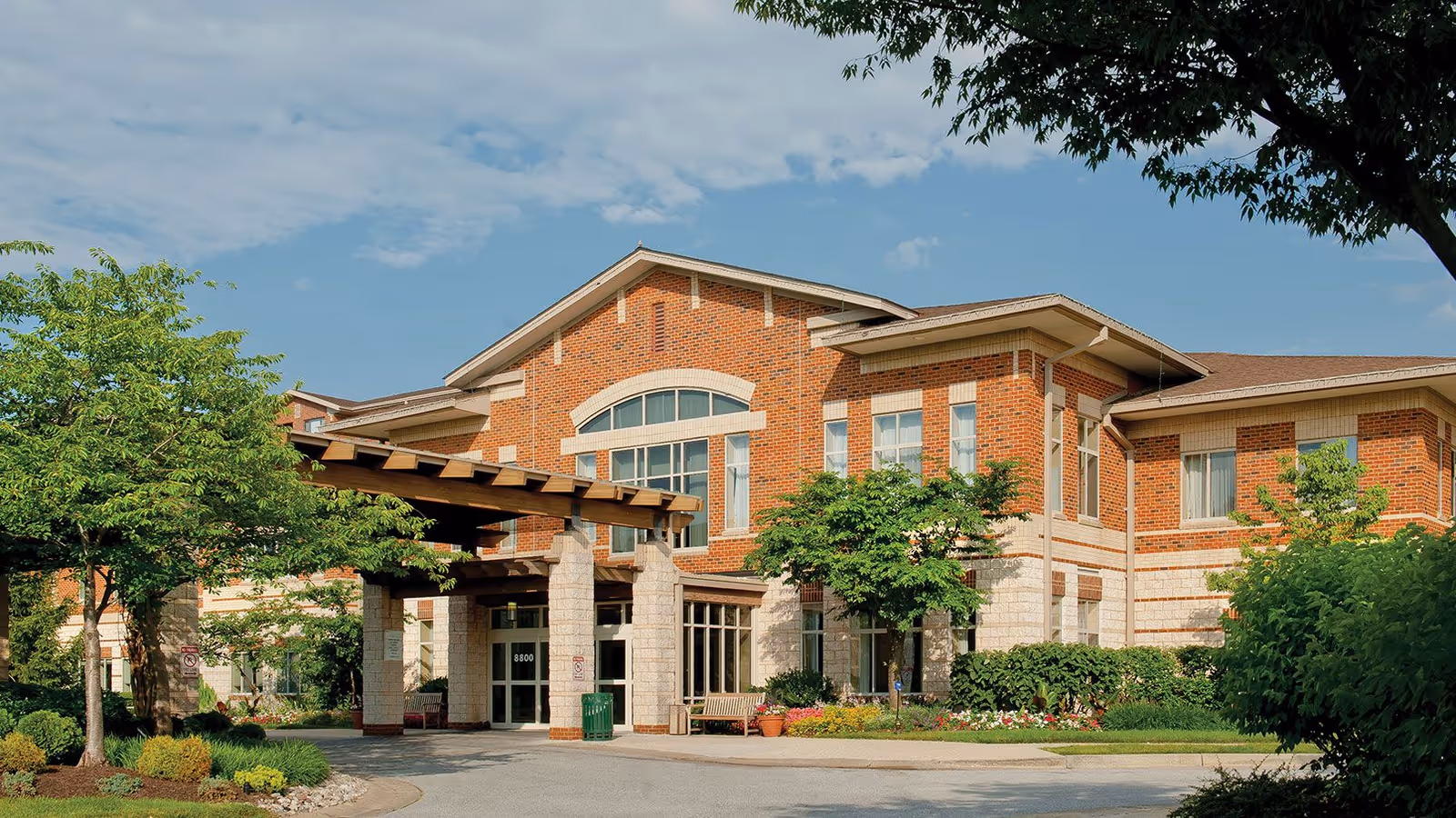Two-story brick senior living building front with a covered entrance, trees, and a landscaped driveway.