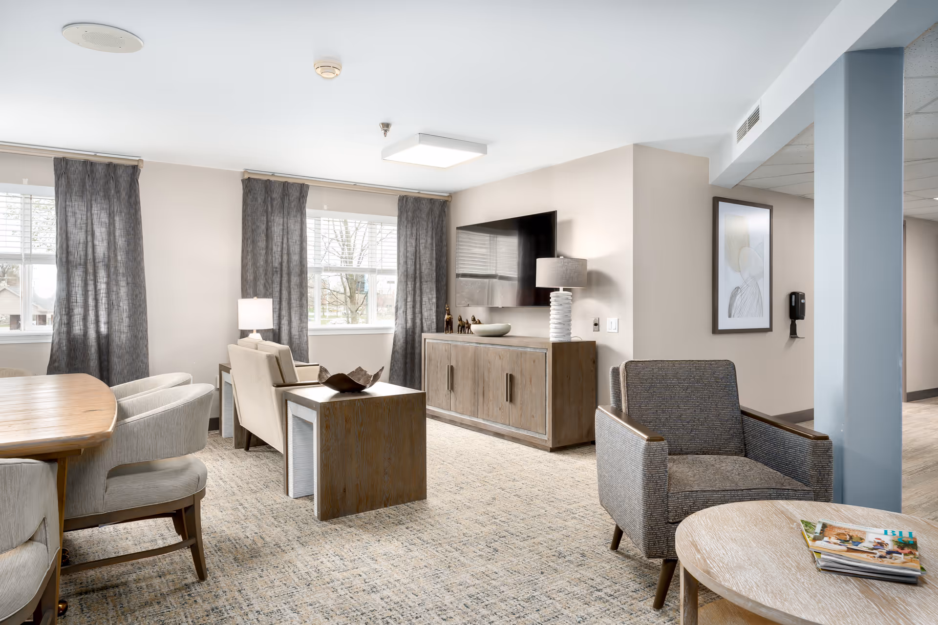 A bright and modern living room area with large windows covered by dark curtains, a wall-mounted flat screen TV above a wooden cabinet, a table lamp, two beige armchairs with a small wooden table between them, a gray armchair, and a round wooden coffee table with magazines on it. The room has light-colored walls and carpeted flooring.