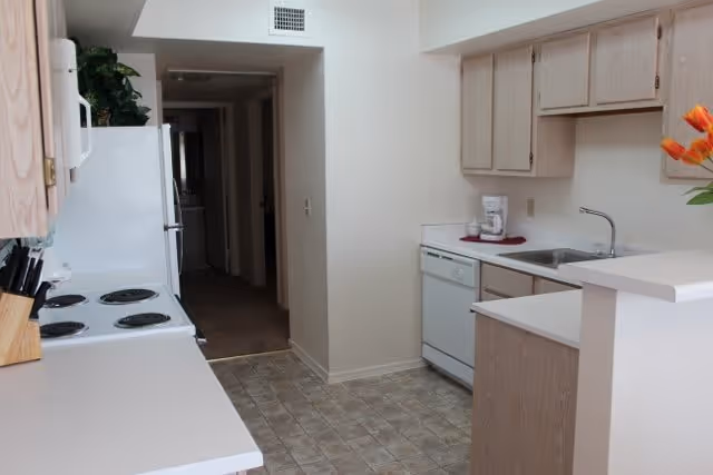 A narrow kitchen with light wood cabinets, white countertops, an electric stove with four burners, a white refrigerator, a dishwasher, a sink, and a coffee maker on the counter. There is a small raised counter area with a vase of orange flowers. The floor is covered with patterned linoleum tiles, and a hallway is visible in the background.