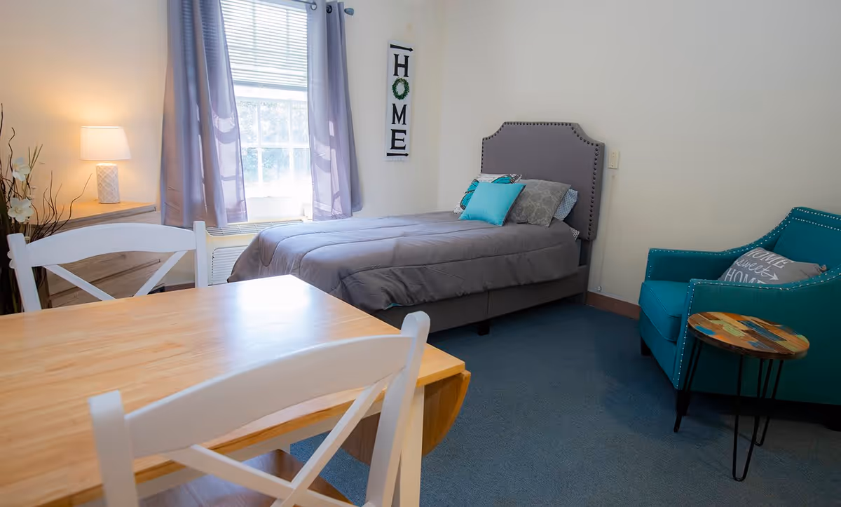 A cozy bedroom with a single bed covered in a gray comforter and several pillows. Next to the bed is a teal armchair with a decorative pillow and a small round wooden side table. A wooden table with white chairs is in the foreground. A window with purple curtains lets in natural light, and a lamp sits on a wooden dresser. A wall hanging with the word 'HOME' is displayed near the bed.