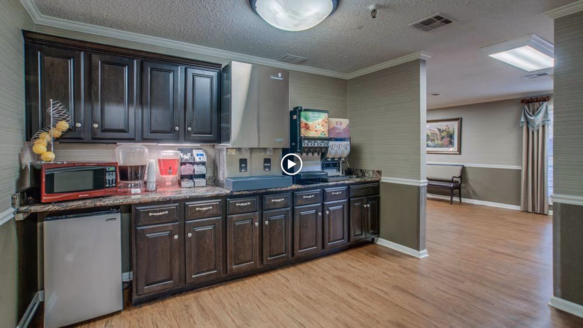 Small kitchenette with dark wood cabinets, countertop appliances (microwave, beverage dispensers, soda fountain) and a mini fridge opening onto a seating area with wood-look flooring.