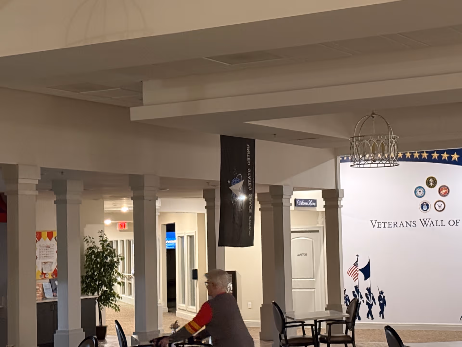 Interior view of a senior living facility hallway with white columns and a Veterans Wall of Honor display on the right wall. A senior person is walking with a walker near a table and chairs. There is a black flag hanging from the ceiling and a plant near the hallway entrance.