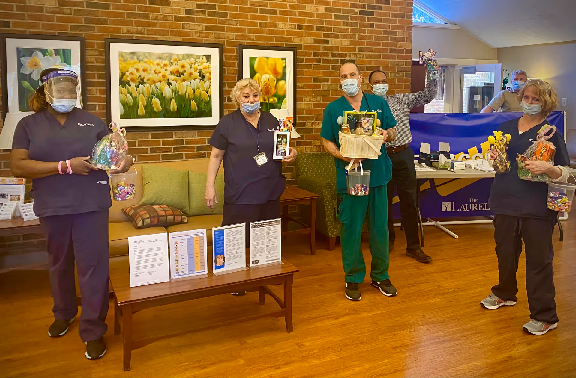 Several masked staff members stand in a senior living facility common room holding gift baskets in front of a brick wall with framed floral pictures.