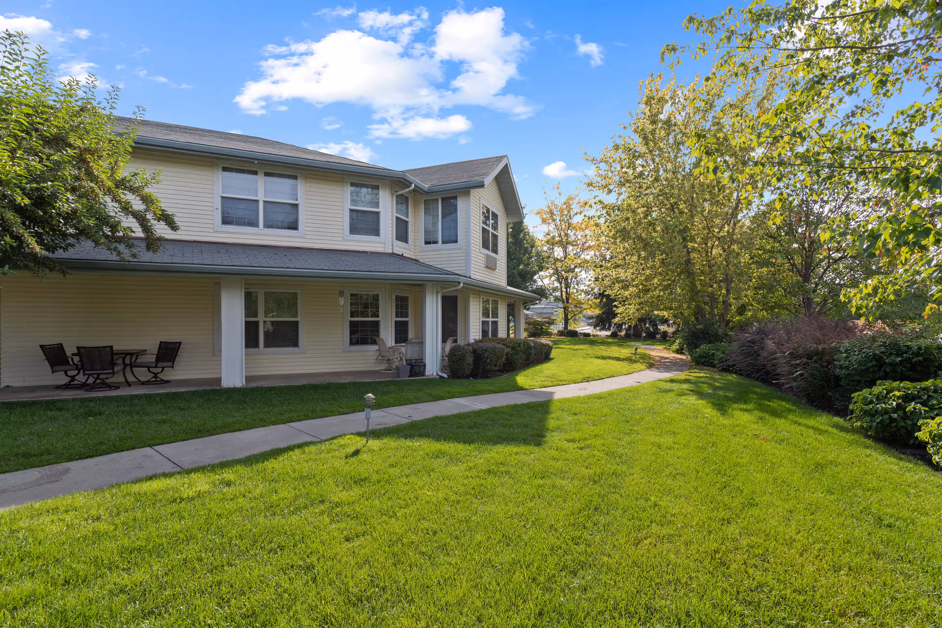 Two-story senior living building exterior with a covered patio, outdoor seating, a curved walkway and well-kept lawn under a blue sky.