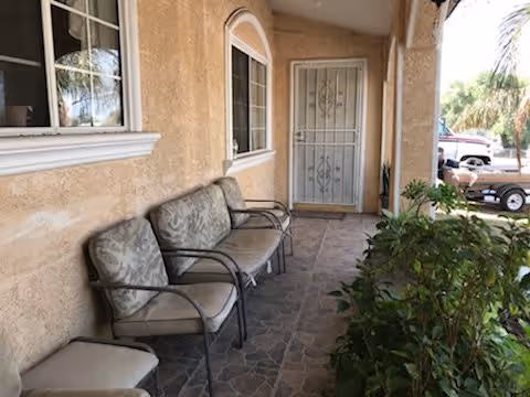 Covered outdoor patio area with three cushioned chairs lined up against a beige stucco wall, a window with white trim, a metal security door, and green plants on the right side. A parked car is visible in the background.