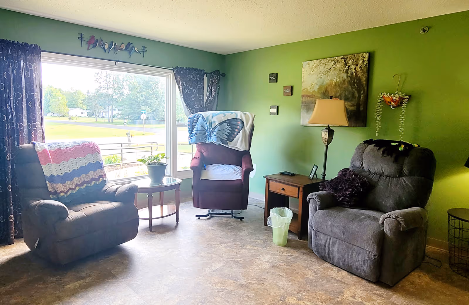 A cozy living room with three recliner chairs, one with a colorful crocheted blanket, another with a butterfly blanket, and the third with a dark purple pillow. A small round table with a potted plant is placed near a large window with dark curtains. The walls are painted green and decorated with a painting, a wall lamp, and a small hanging basket with flowers. A wooden side table with a lamp and a small trash bin are also visible.
