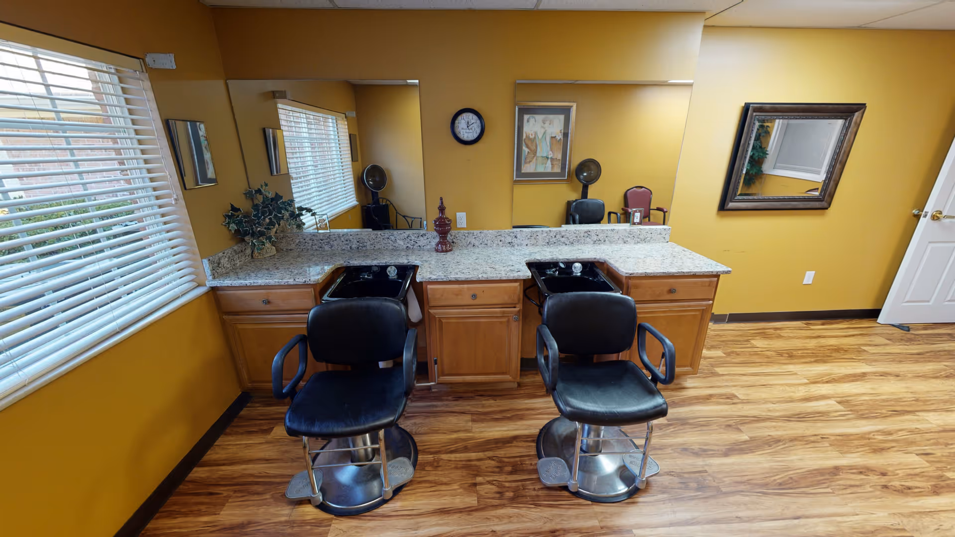 A salon area with two black styling chairs in front of wash sinks and mirrors, a granite countertop, and wood flooring.