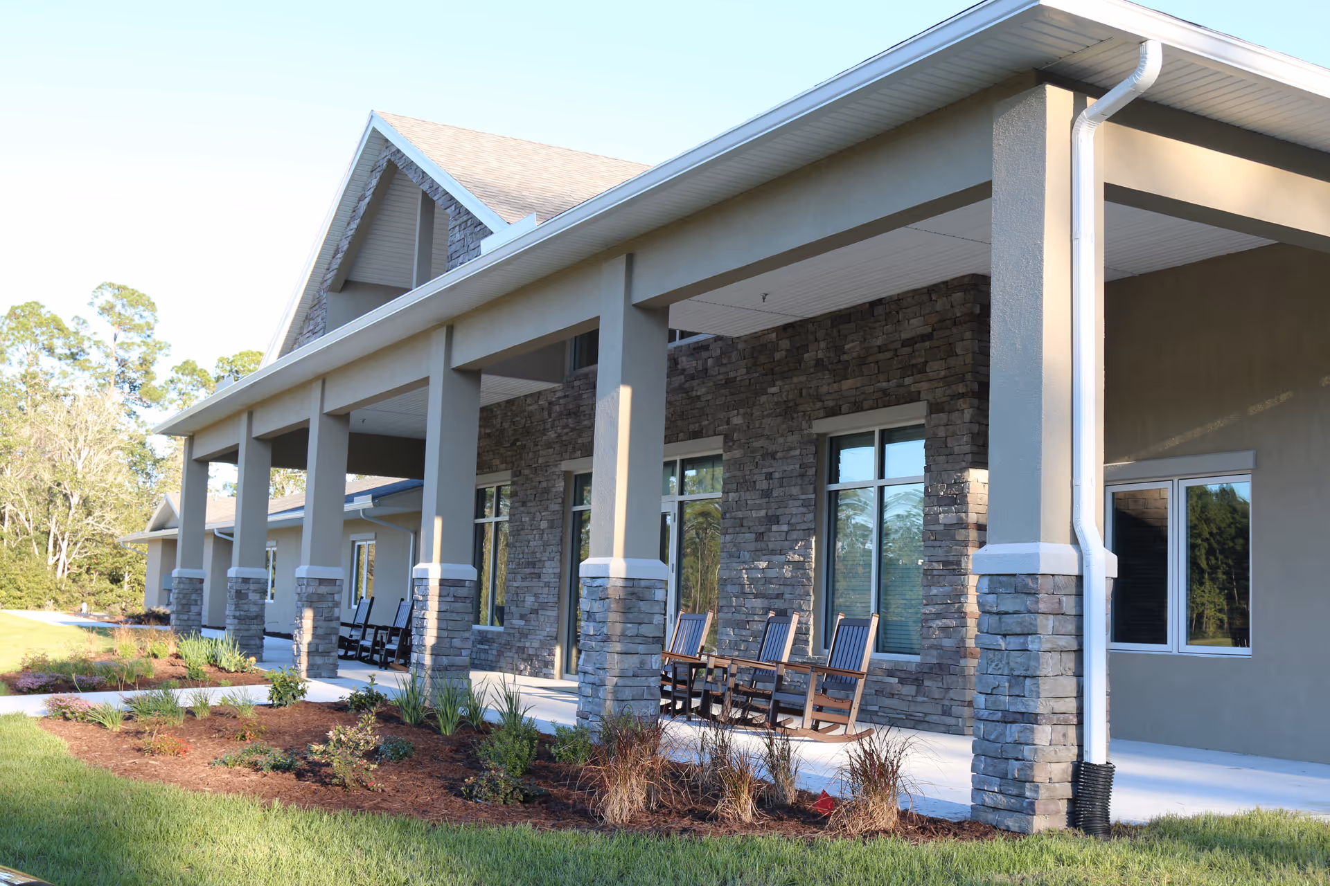 Exterior view of a senior living facility with a covered porch supported by stone and concrete columns. Several wooden rocking chairs are placed on the porch. The building has large windows and a landscaped garden with grass and small plants in front.