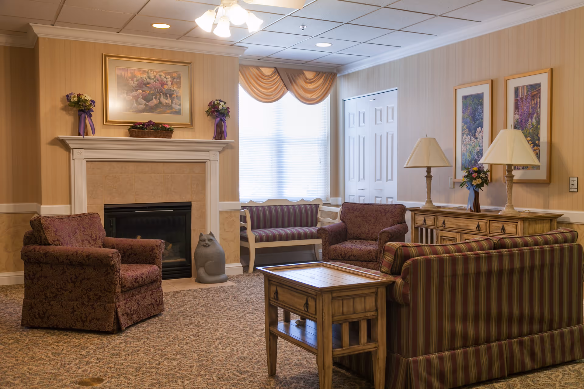 A cozy living room area in a senior living facility with patterned armchairs and a striped sofa arranged around a wooden coffee table. There is a fireplace with a framed floral painting above it, flanked by two flower arrangements. A window with sheer curtains and a valance lets in natural light. A wooden sideboard with two lamps and floral decorations is against the wall, which is adorned with two framed floral pictures.