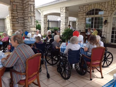 Several elderly residents, many in wheelchairs, seated at tables under a covered stone patio eating and socializing.