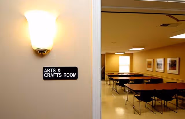 View of an arts and crafts room with several tables and chairs arranged in rows. The room has beige walls, framed pictures on the wall, and a window letting in natural light. A wall-mounted light fixture and a sign reading 'ARTS & CRAFTS ROOM' are visible near the entrance.