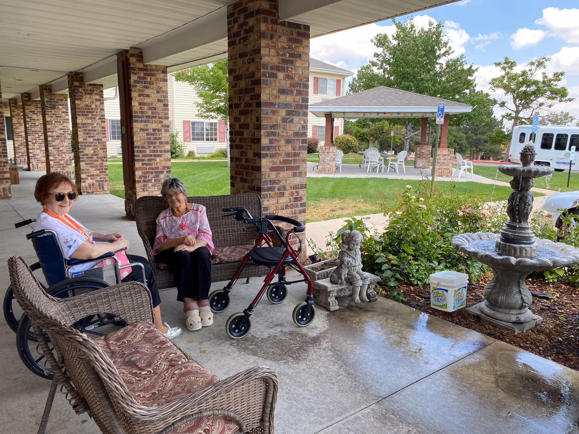 Two elderly women sitting and smiling on a covered patio area with wicker furniture and a walker. One woman is in a wheelchair wearing sunglasses, and the other is seated on a wicker loveseat. There is a decorative stone fountain and a small statue of a girl sitting on a bench nearby. In the background, there is a grassy area with a gazebo, white chairs, and a white van parked near a building with red shutters.
