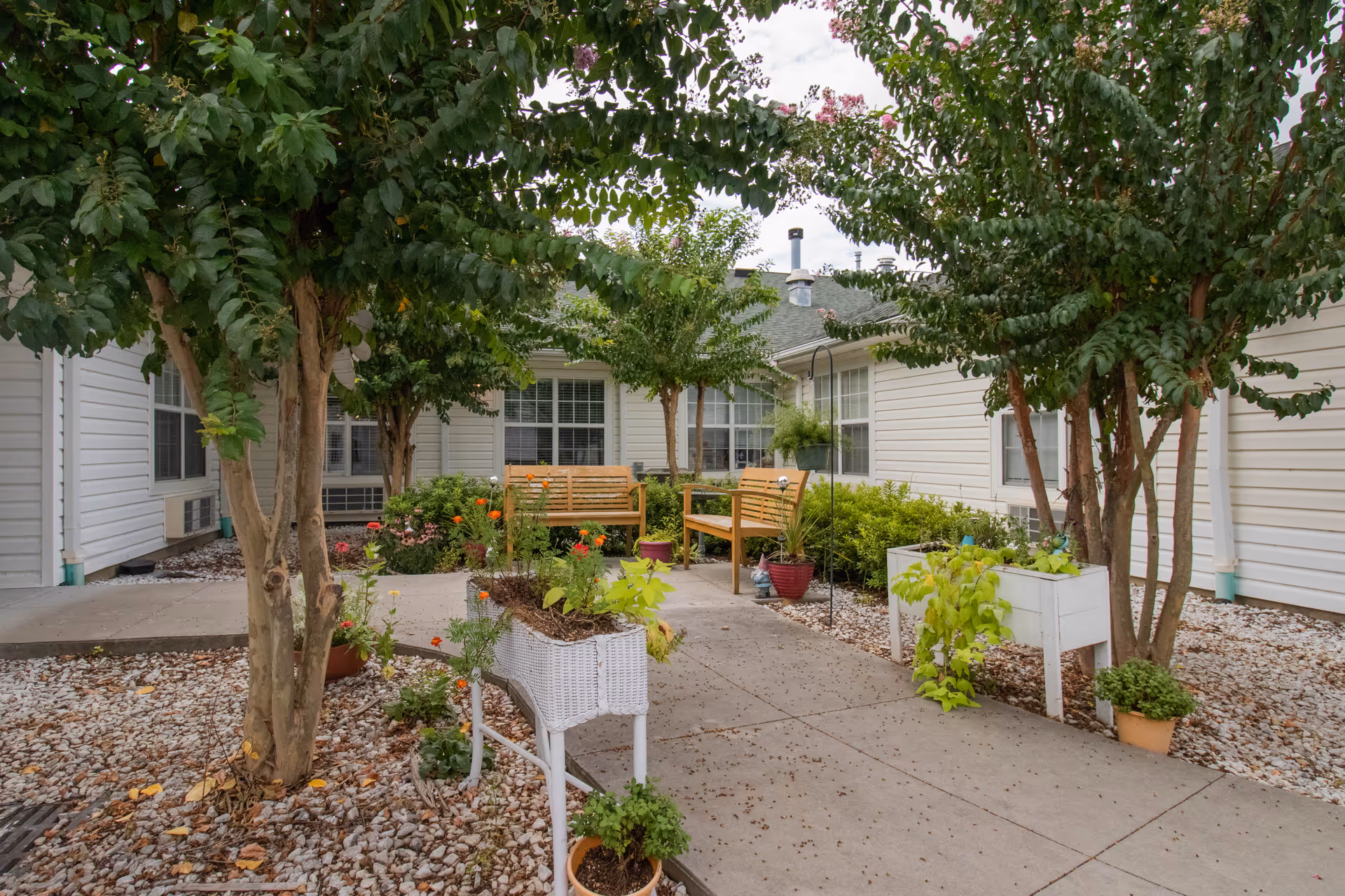 Outdoor courtyard area at Pee Dee Gardens featuring a paved walkway, two wooden benches, several trees, and various potted plants and flowers arranged around the space. The courtyard is surrounded by the building's white exterior walls and windows.