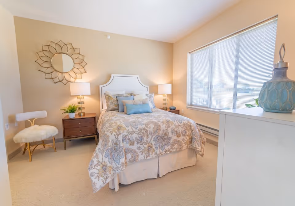 A bright and cozy bedroom with a bed featuring a floral patterned comforter and multiple pillows. There are two bedside tables with lamps on each side of the bed. A decorative round mirror hangs on the wall above one bedside table, which also holds a small potted plant. A white cushioned chair with gold legs is positioned near the wall. A large window with blinds allows natural light to fill the room. A white dresser with a decorative blue vase is visible in the foreground.